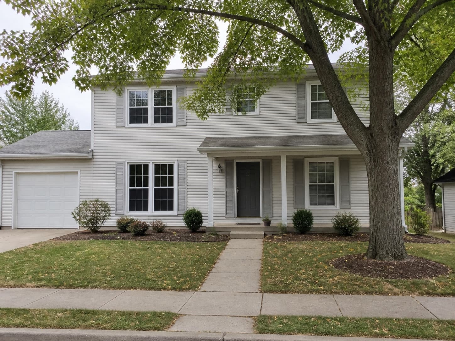 Illustrative original view of a modest light-gray suburban home with a simple foundation bed and front walkway.